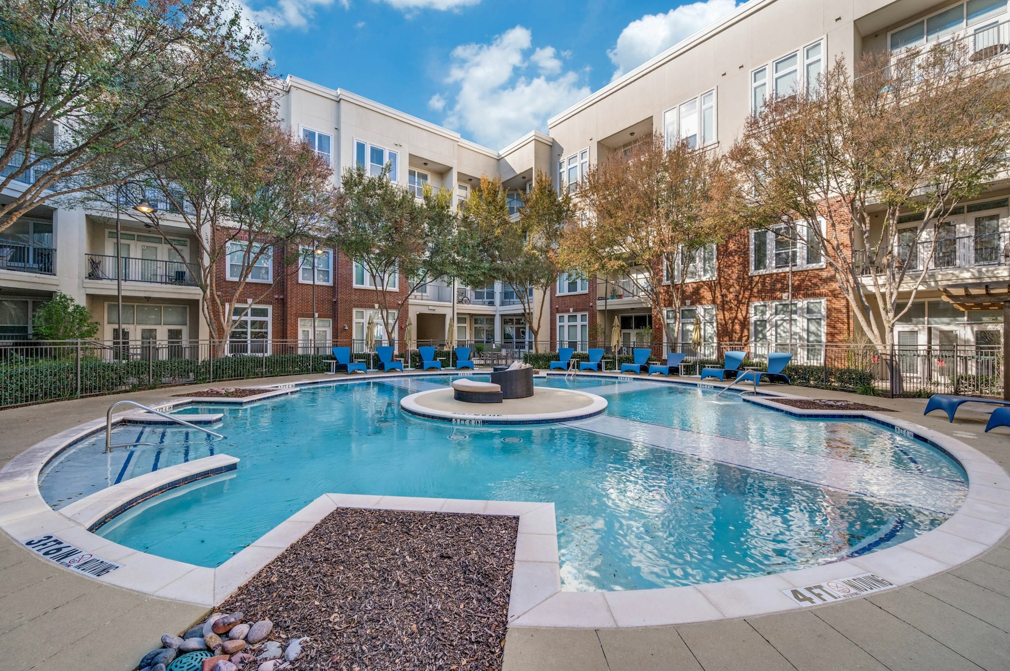 AMLI Frisco Crossing - Courtyard view of round resort-style pool with blue lounge chairs, trees, and red brick buildings at AMLI Frisco Crossing apartments