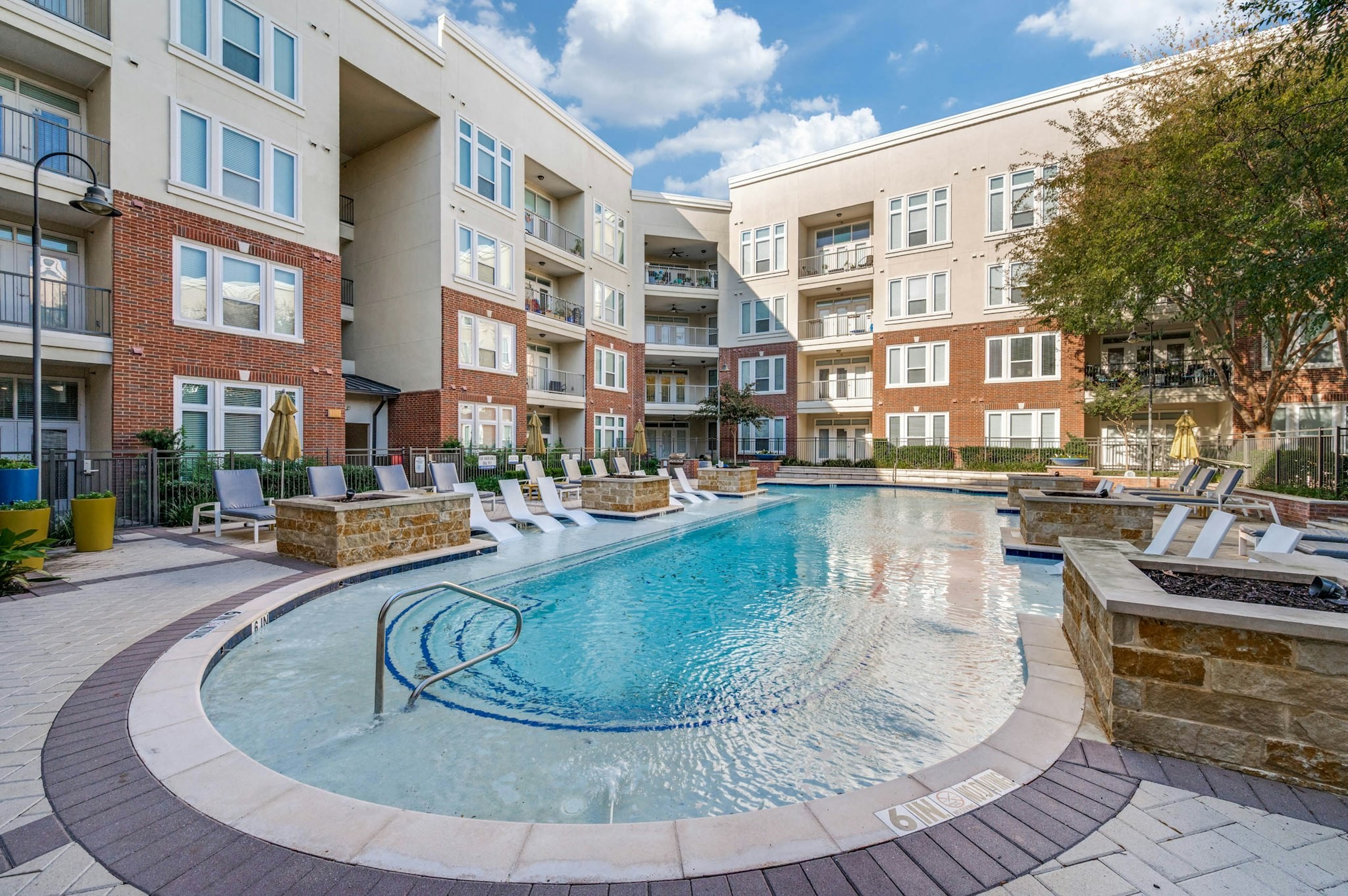 AMLI Frisco Crossing - AMLI Frisco Crossing apartment daytime pool view with tanning ledge, lounge seating, and surrounding red brick buildings and trees