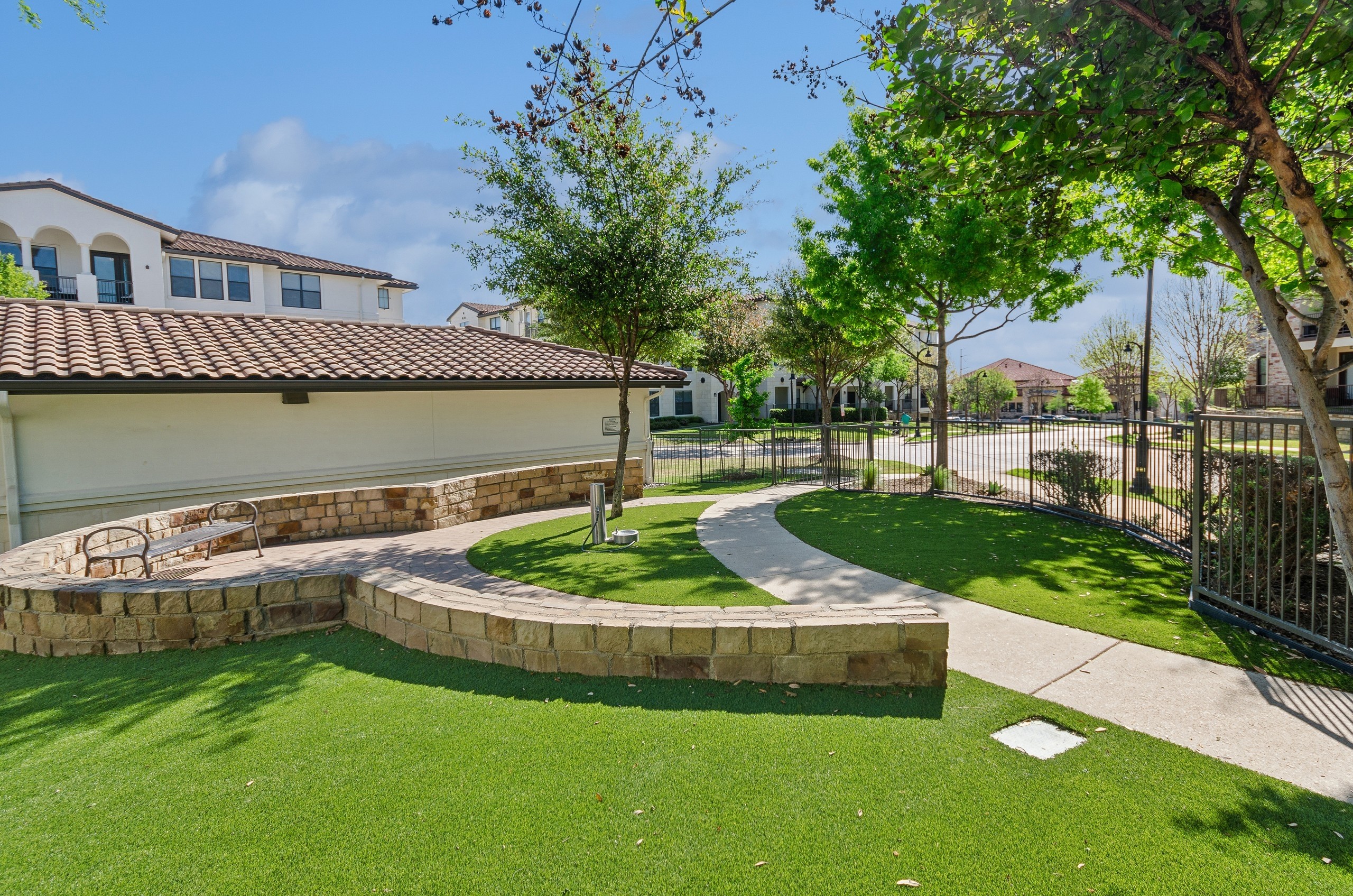 A fenced dog park at AMLI on Riverside featuring green turf and a curved stone wall surrounding a bench and a paved pathway