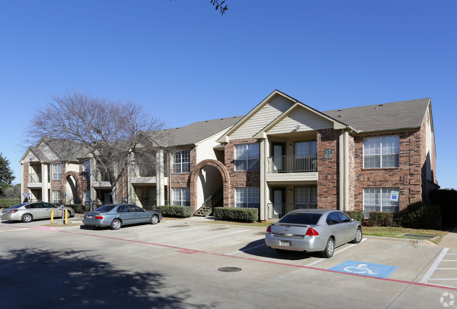 A parking lot in front of a brick apartment building with a car parked in the handicap spot. at Ash Lane Apartment Homes