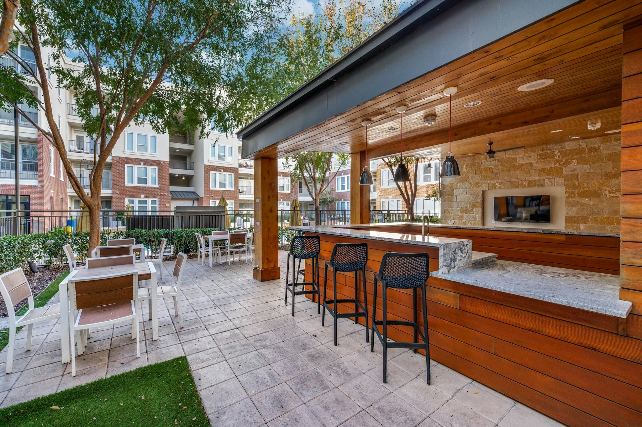 AMLI Frisco Crossing - Covered outdoor lounge with bar seating, pendant lights and flat screen TV at AMLI Frisco Crossing apartments, with tables and trees nearby